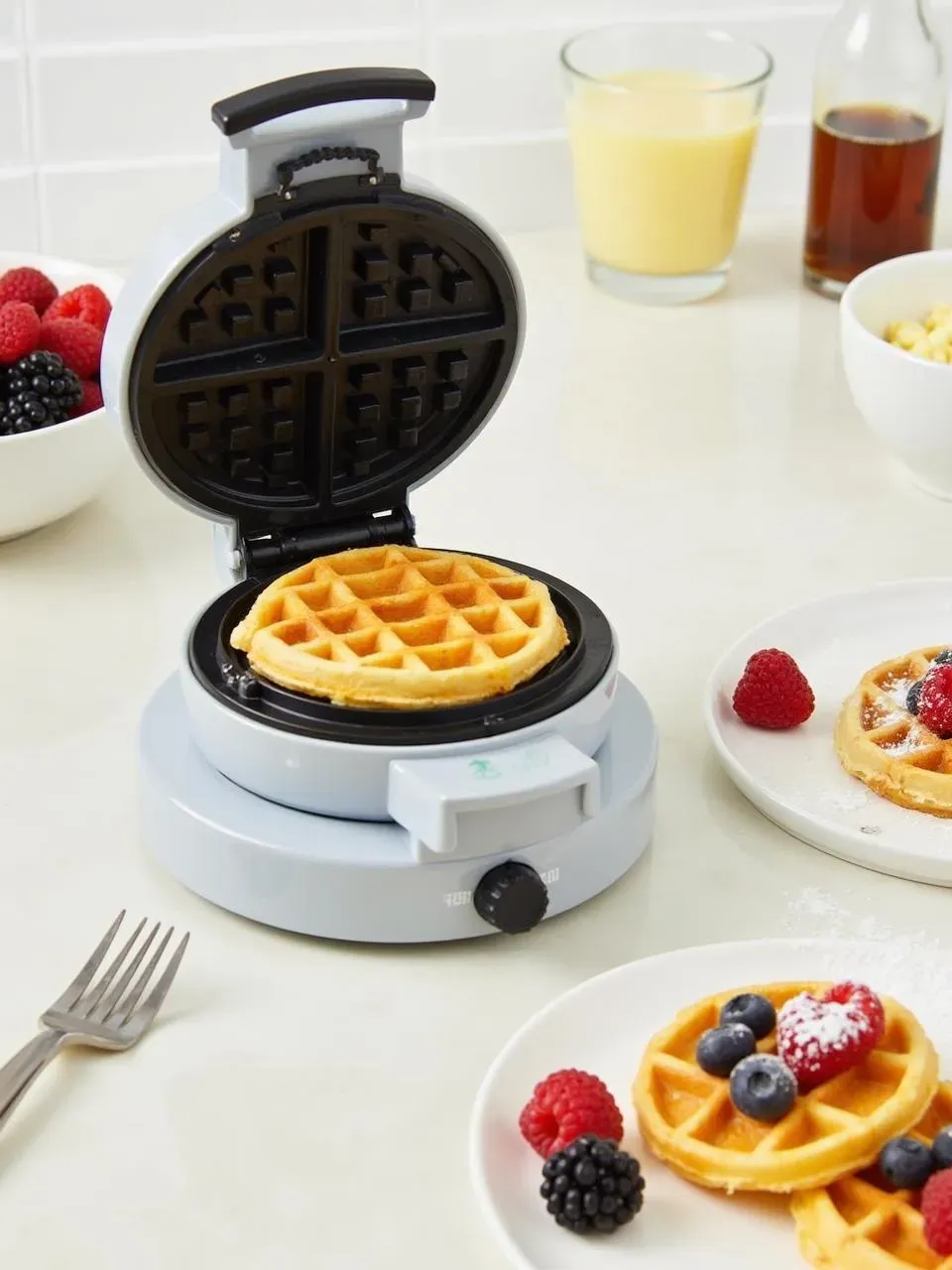 Dash Mini Waffle Maker cooking a waffle with berries, syrup, and finished waffles on a plate in a cheerful kitchen setting.