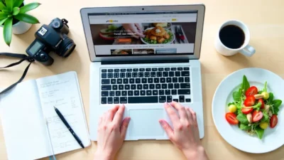 Top view of a food blogger’s workspace with a laptop, recipe notes, camera, and a plate of styled food on a clean surface.