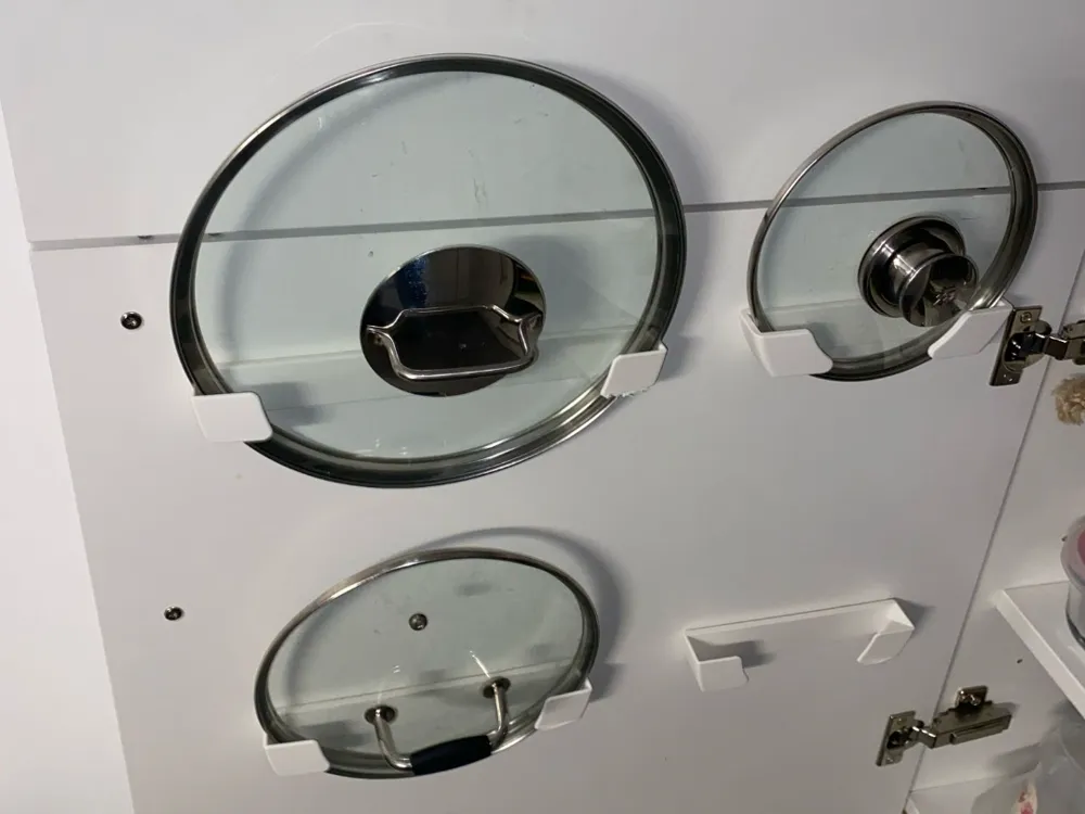 Three glass pot lids stored vertically using white 3D printed holders mounted on the inside of a kitchen cabinet door for organized and space-saving storage