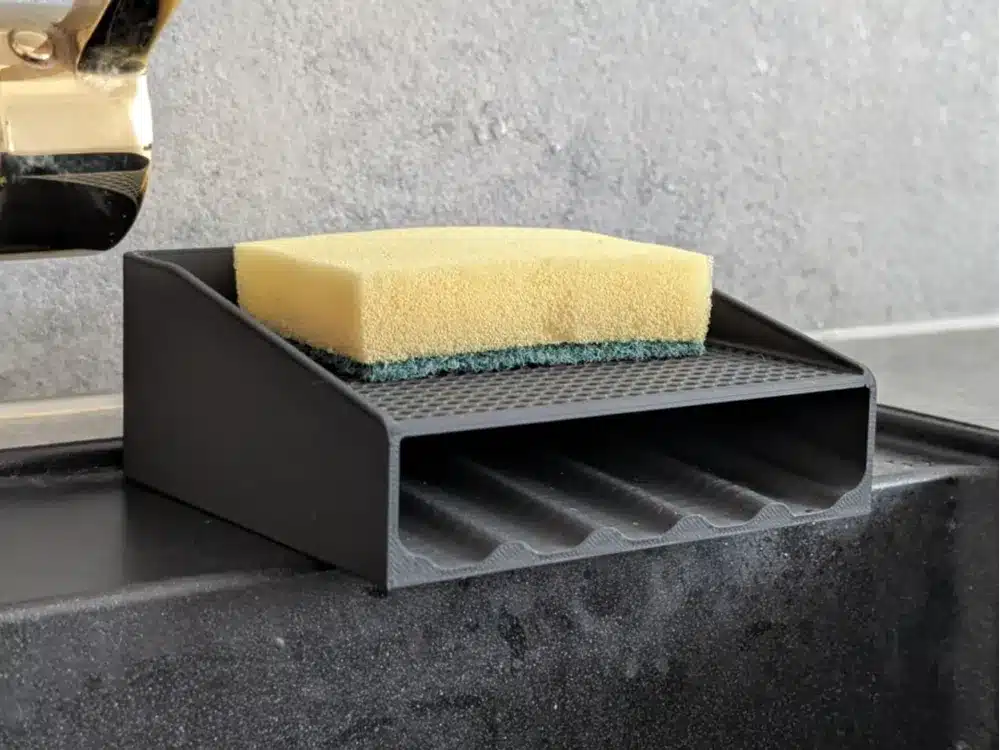 Black 3D printed sponge holder placed on a kitchen sink, featuring angled grooves and a raised surface for draining and keeping sponges dry