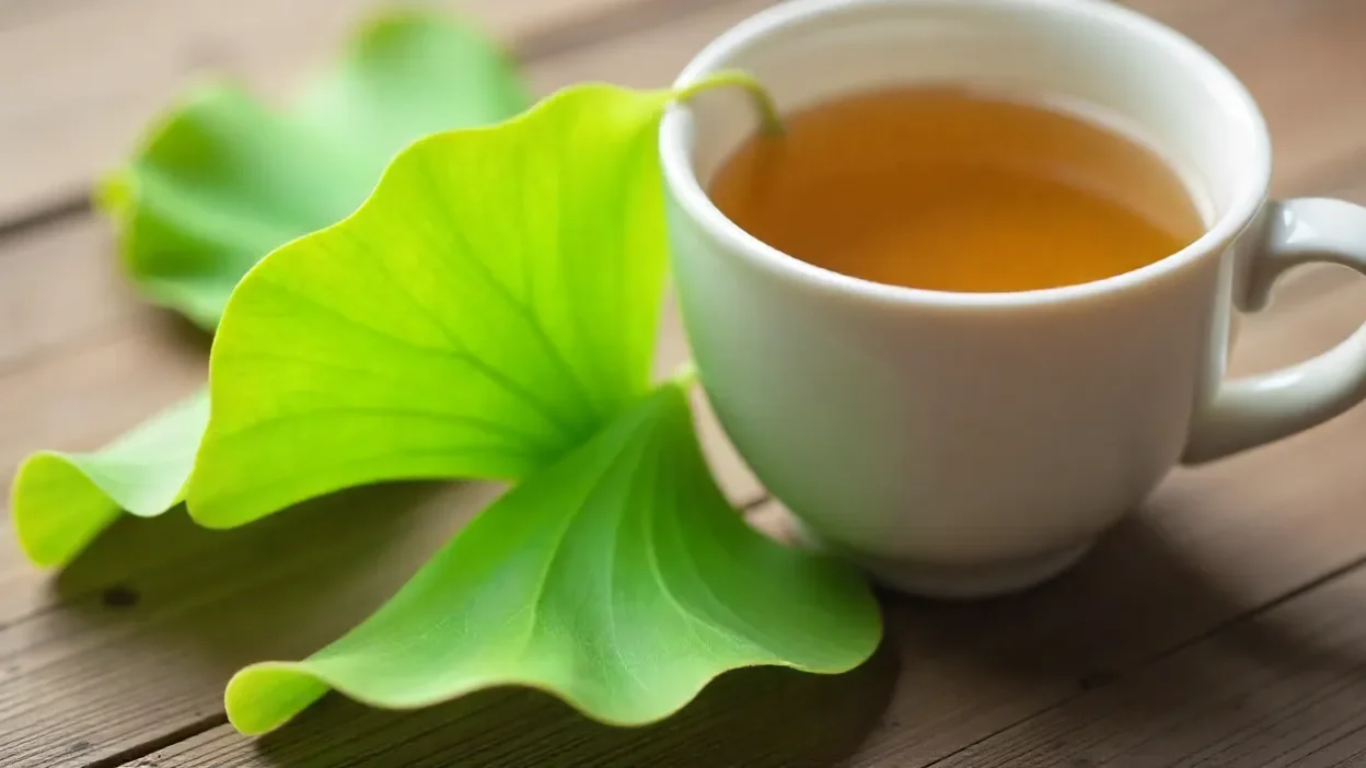 Fresh ginkgo biloba leaves next to a cup of herbal tea on a wooden surface in soft natural light.